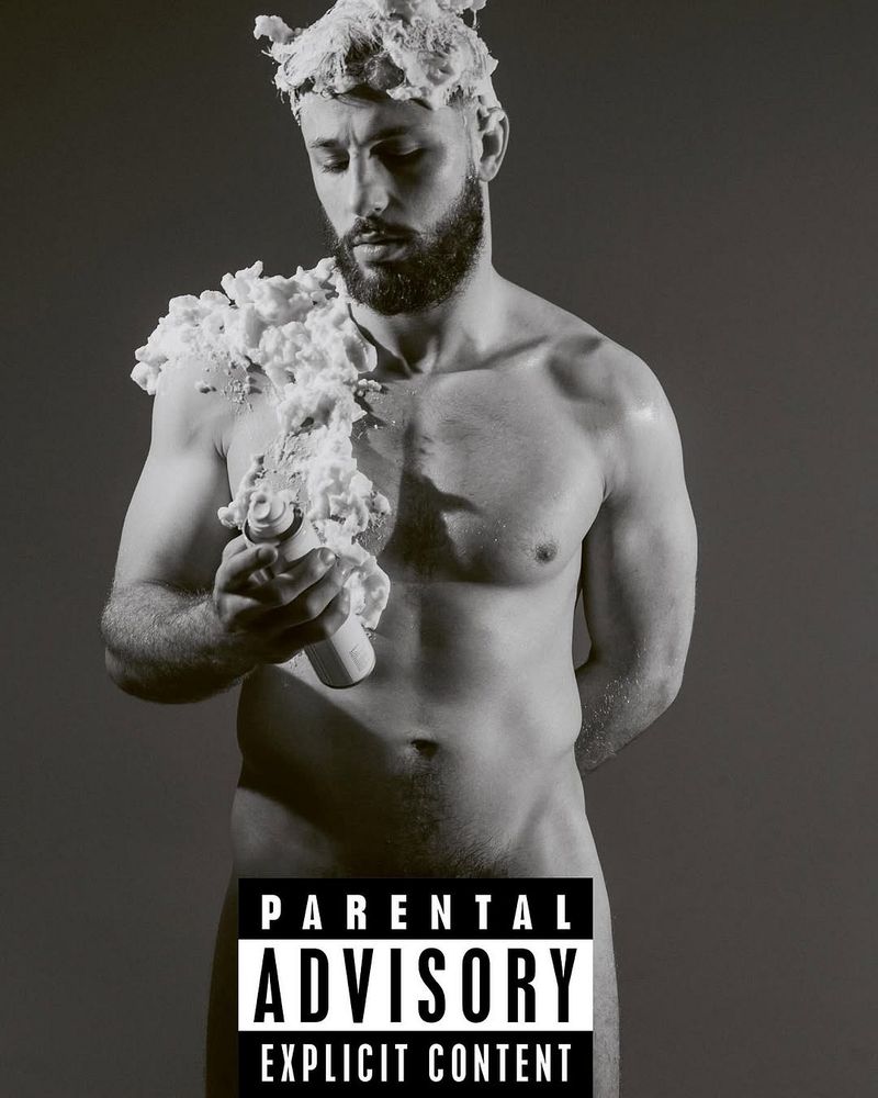 Monochrome portrait of a shirtless adult man with shaving cream on his head and shoulder, holding a can, against a grey backdrop.