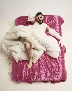 Man wearing white dress and sunglasses lying on a magenta satin bed cover
