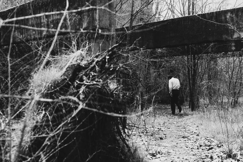 Monochrome image of a man walking on a path under a concrete structure in a forest with tangled branches in the foreground.