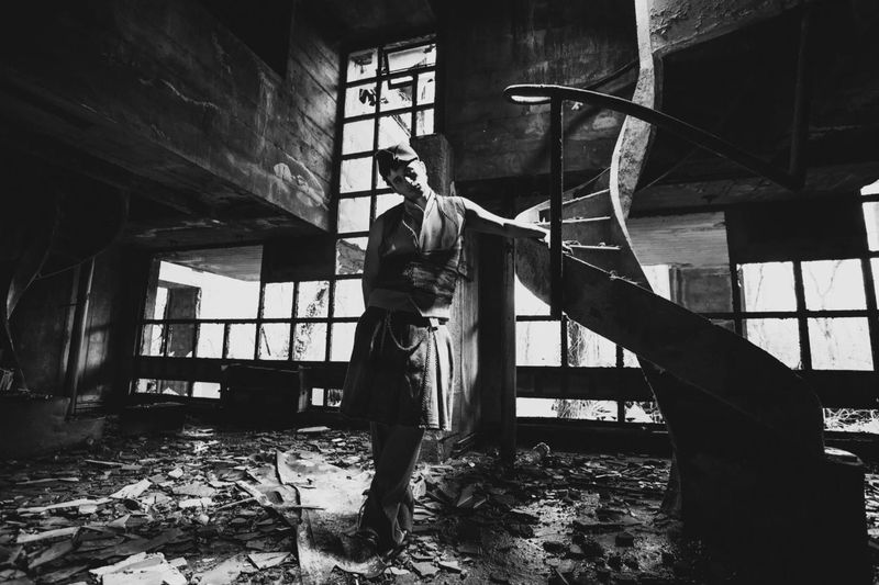 Monochrome portrait of a person in a vest and cap standing in a dilapidated interior with a spiral staircase and broken windows.