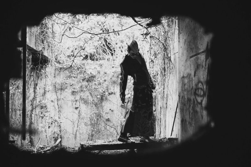 Person in flowing skirt and hooded top stands inside dilapidated structure covered with vines in artistic black and white photograph.
