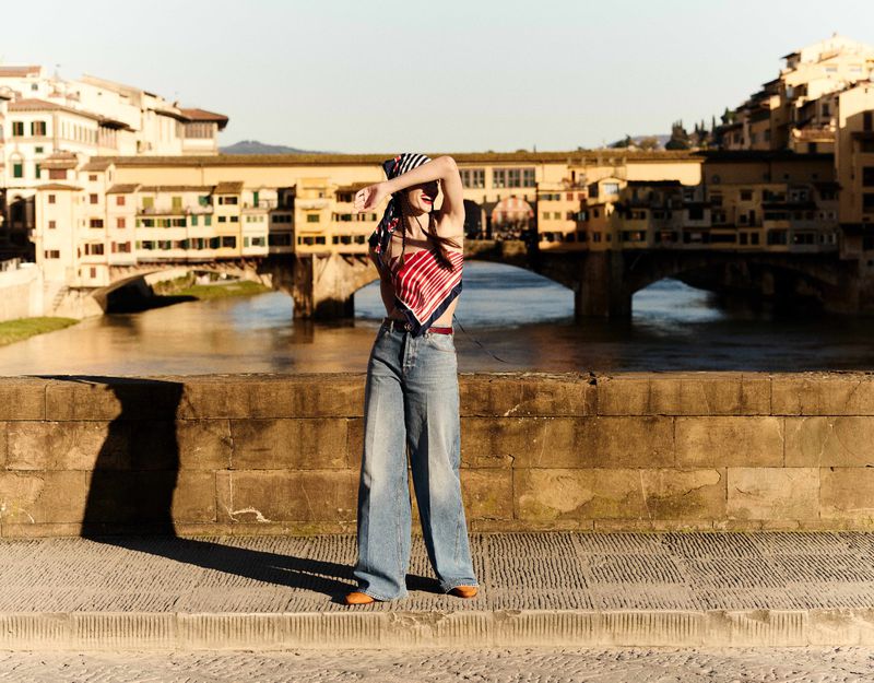 A woman poses on a stone bridge wearing jeans and a patterned headscarf in Florence with the Ponte Vecchio visible in the background.