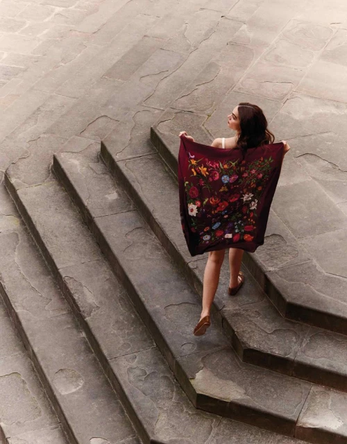 Woman in stylish floral scarf ascends stone steps outdoors in a full shot, showcasing fashion and architectural backdrop with natural light.