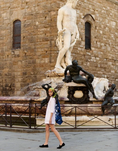 Fashion shot in Italy: adult woman in a white dress walks past a fountain and classical sculpture of Neptune, in a sunny outdoor setting.