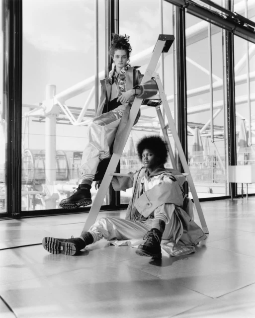 Two fashion models in matching silvery outfits pose with a metal ladder against a modern window backdrop with a geometric metal structure.
