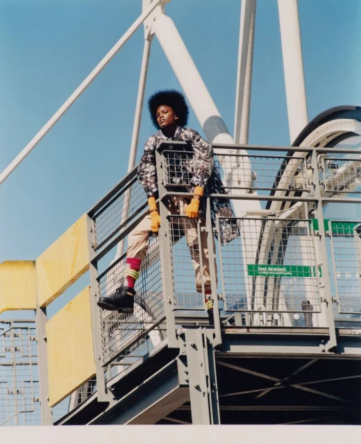 A woman poses on an industrial metal structure against a light blue sky, wearing a patterned jacket and orange gloves in a fashion shot.