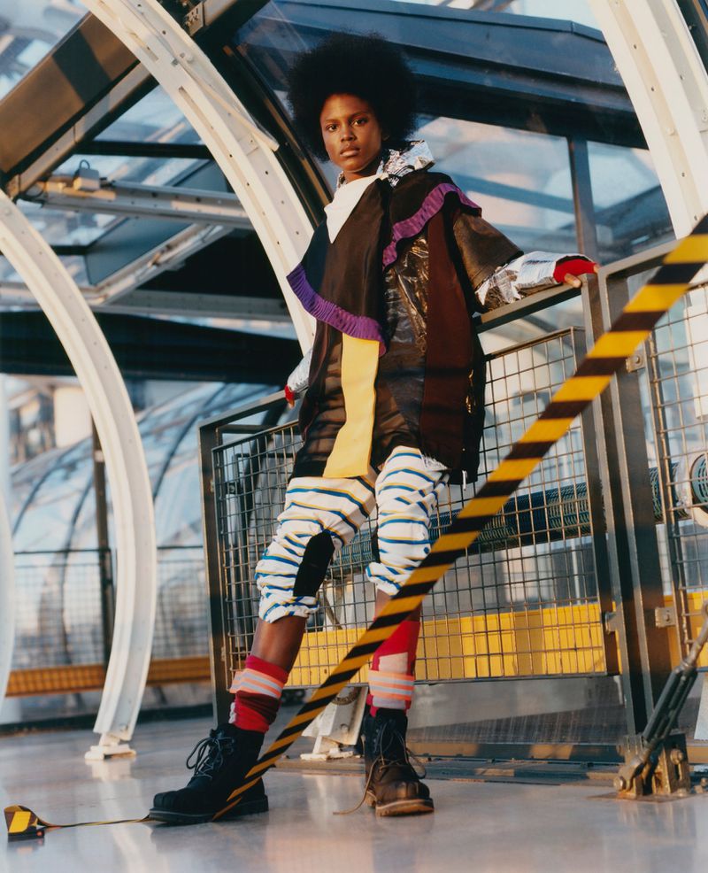 Fashion editorial shot of a Black female model in avant-garde outfit, posing on modern staircase with natural light and striped barrier tape.