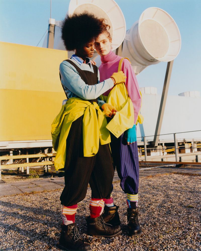 Two fashion models on a rooftop in brightly colored clothing pose closely together in front of ventilation shafts and yellow walls.