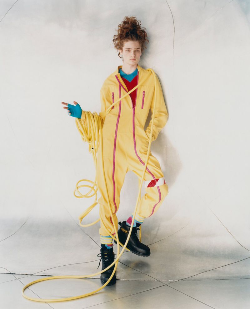 Full-shot of a person in a yellow jumpsuit with a red top, black boots, and curly red hair, standing on a reflective surface indoors.