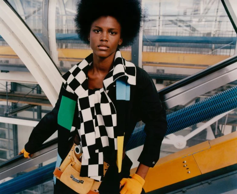 Stylish adult with afro stands next to escalator wearing black jacket with checkered scarf, tool belt and orange gloves in transit hub.