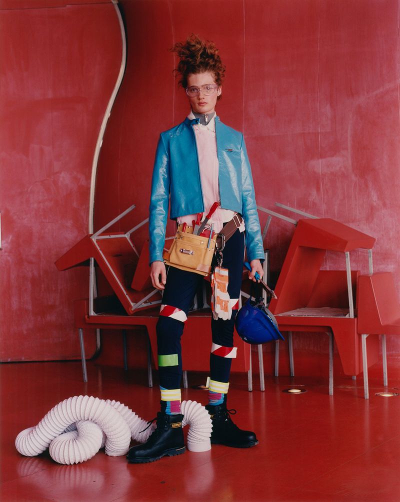 Person in a blue jacket poses with a toolbox and work helmet against a red backdrop with stacked chairs and a white tube.