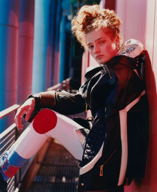 Model with curly hair poses outdoors in futuristic fashion: black jacket, patterned leggings, and metallic shoulder wrap in urban setting.