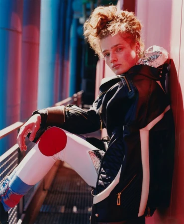 Model with curly hair poses outdoors in futuristic fashion: black jacket, patterned leggings, and metallic shoulder wrap in urban setting.