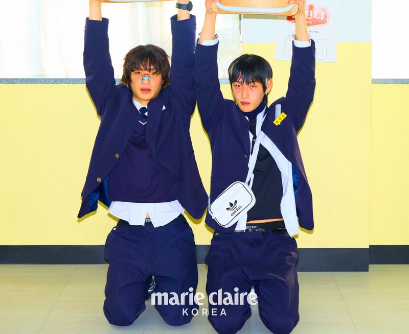 Two young men in school uniforms kneel holding chairs overhead against a yellow wall in an indoor setting, as featured in Marie Claire Korea.