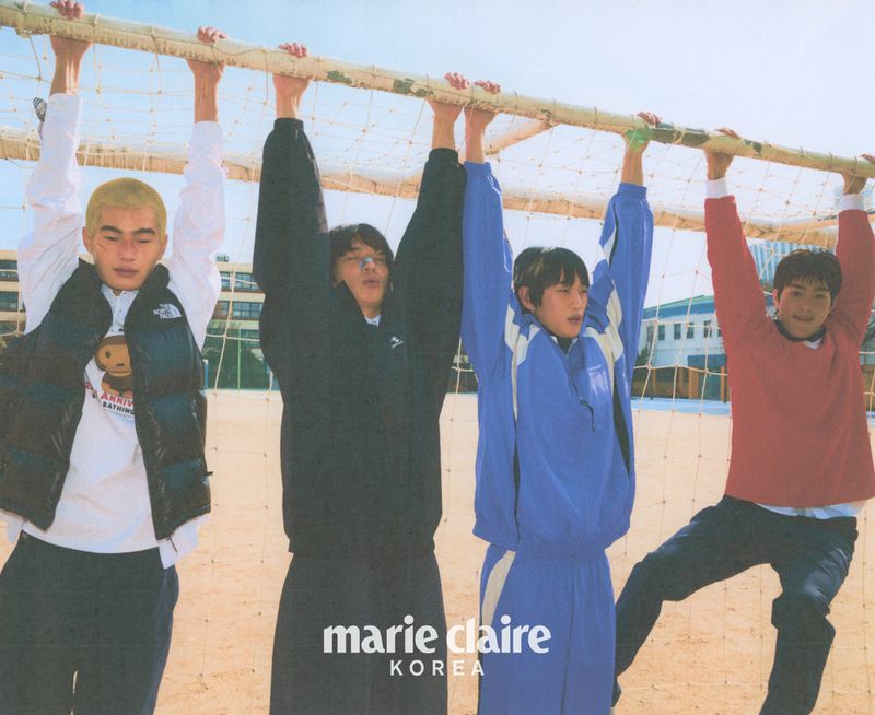Four young adult men hanging from a soccer goal post net, wearing sportswear, photographed outdoors under a bright sky.
