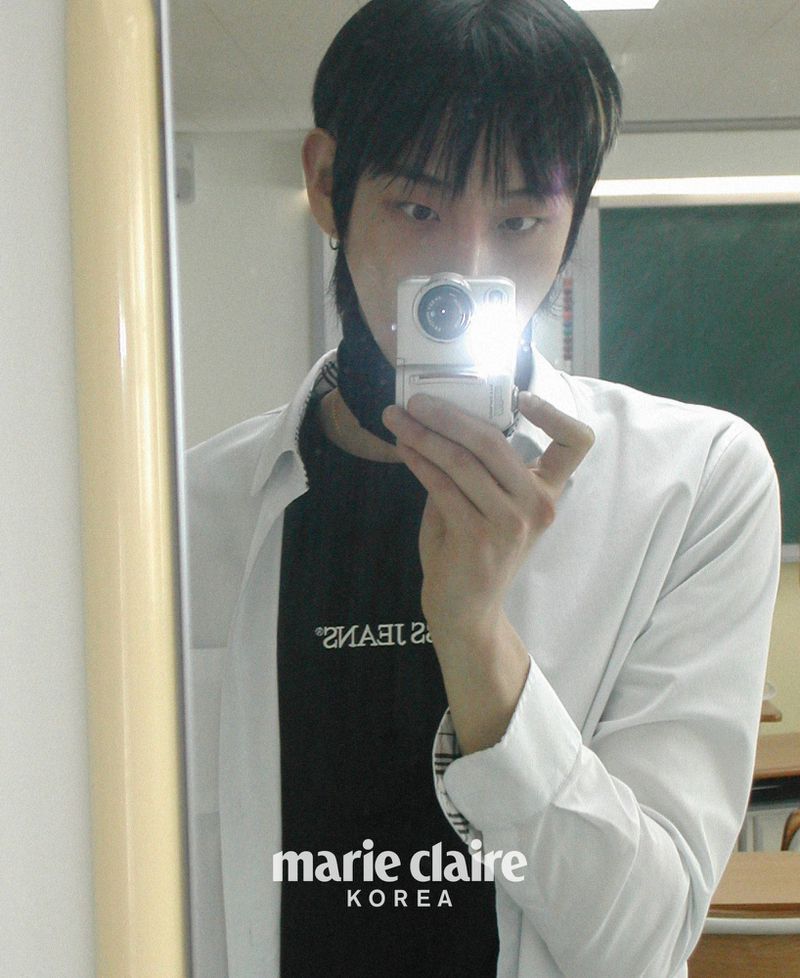 Adult male takes a selfie in a classroom setting. He wears a black shirt under a white button-up and holds a digital camera up to a mirror.