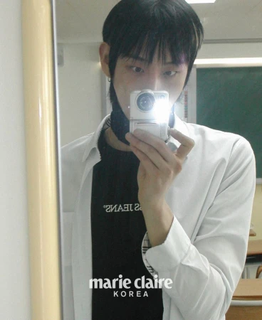Adult male takes a selfie in a classroom setting. He wears a black shirt under a white button-up and holds a digital camera up to a mirror.
