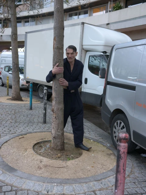 A man in a black jumpsuit hugs a tree on a cobblestone street next to parked vans, with a light-colored building in the background on a sunny day.