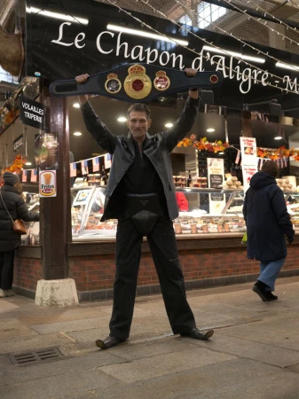 Adult man holding a boxing champion belt over his head in an indoor market with food stalls and shopfronts in the background.