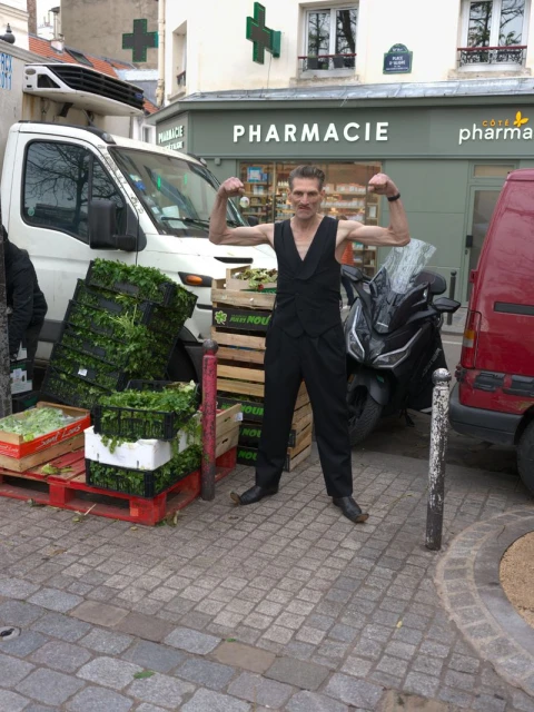 Man flexing biceps on a city street next to a delivery truck with crates of produce. Pharmacy building in the background.