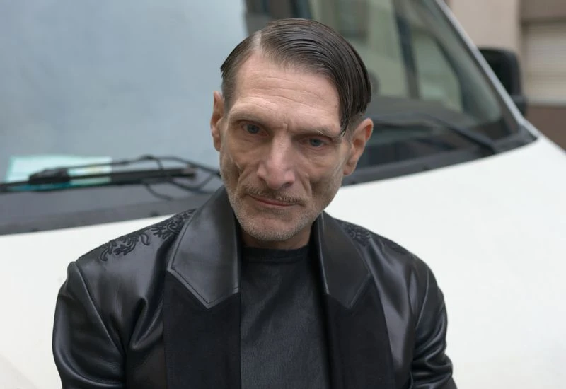 Close-up portrait of a serious adult male with a dark jacket standing outdoors, a white van blurred in the background.