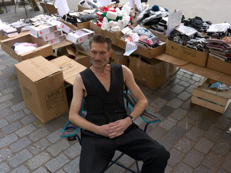 Adult man sitting in a chair outdoors next to tables displaying goods, wearing a vest and dark pants on a cobblestone street.