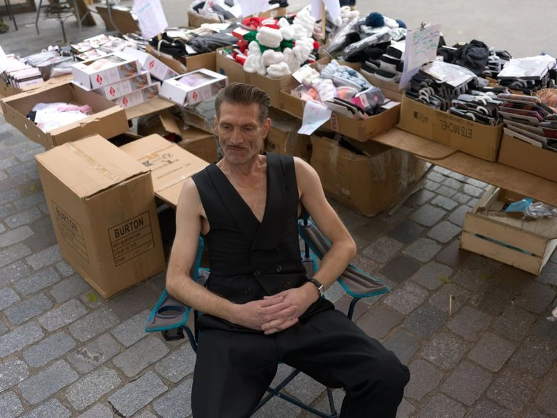 Adult man sitting in a chair outdoors next to tables displaying goods, wearing a vest and dark pants on a cobblestone street.