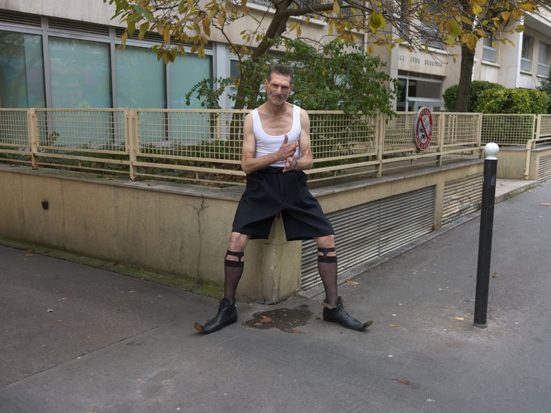 Man in white tank top and dark shorts standing on a city street against a building wall
