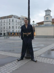 Man wearing a dark cropped blazer and trousers posing outdoors in a public square with buildings and a clock tower in the background.