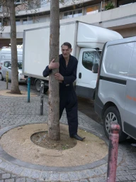 Man wearing a dark jumpsuit hugging a tree trunk on a paved urban street with parked delivery vans in the background.