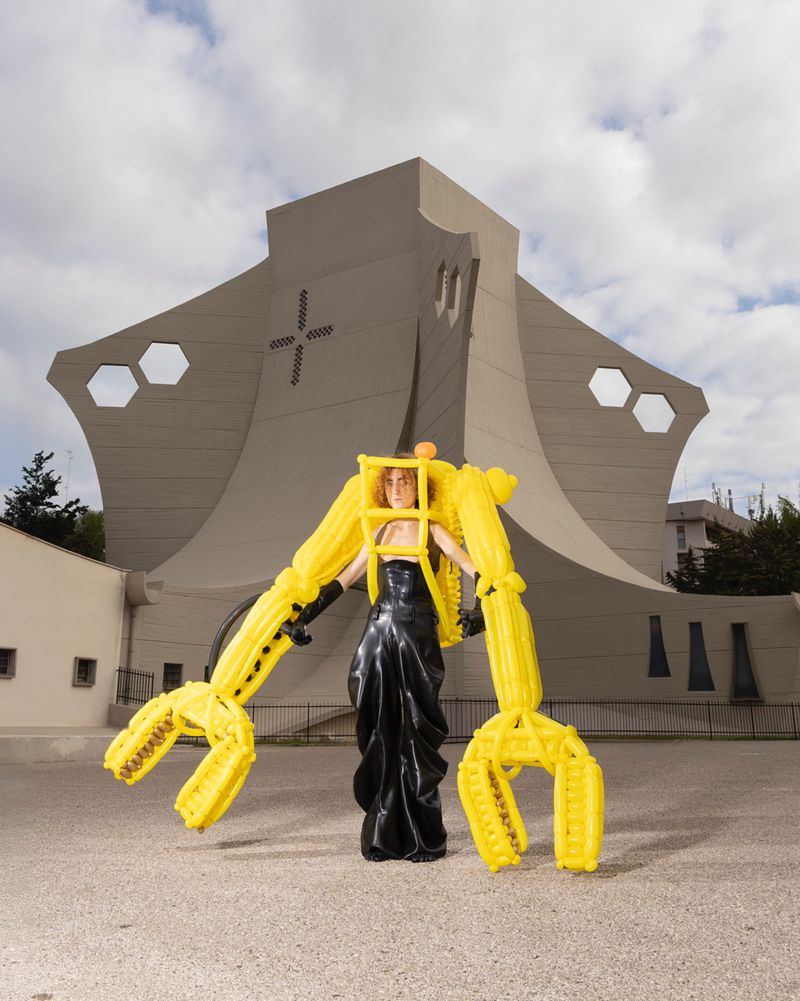 Woman in avant-garde yellow exoskeleton and black gown poses in front of architectural building in an outdoor setting on a cloudy day.