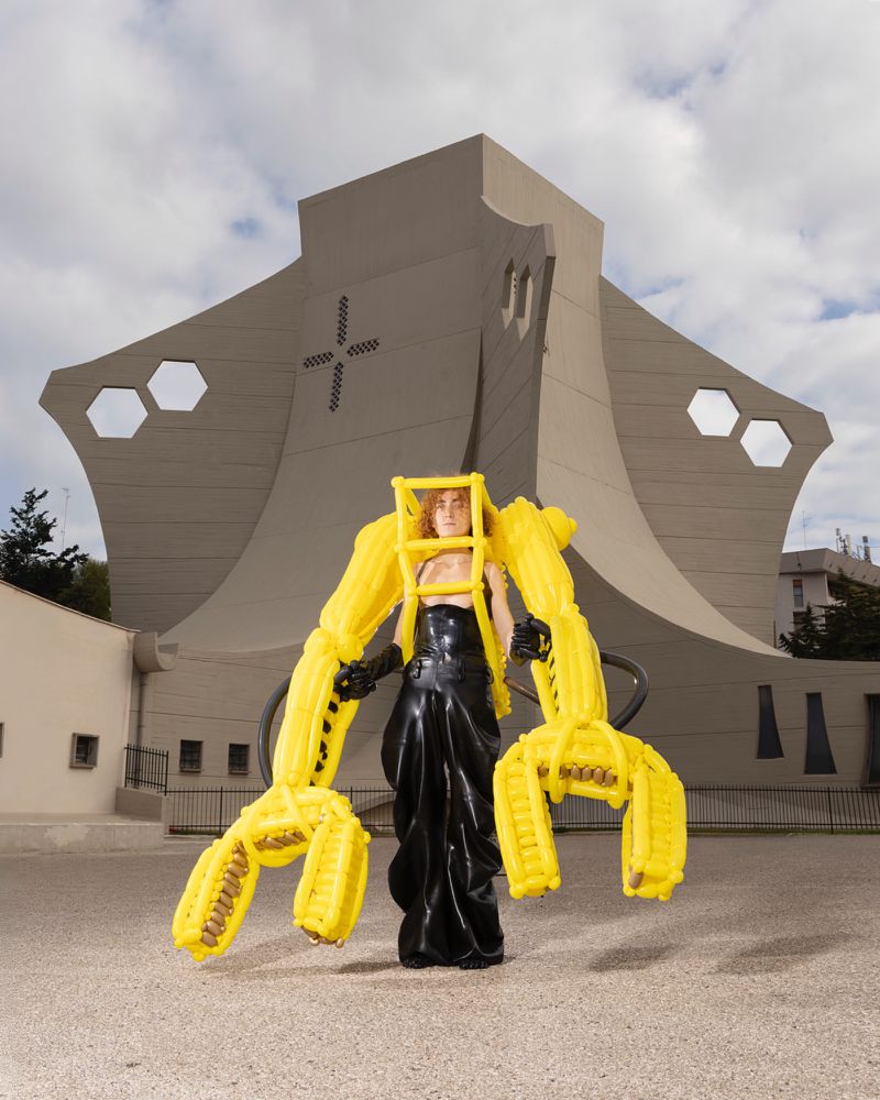 A woman in a black dress and yellow frame stands before a towering concrete building with a unique design under a partly cloudy sky.