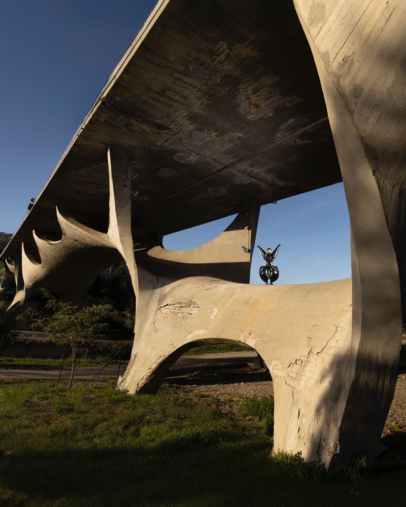 Exterior view of an abstract, concrete sculpture with a butterfly perched on top, set against a clear blue sky and grassy landscape.