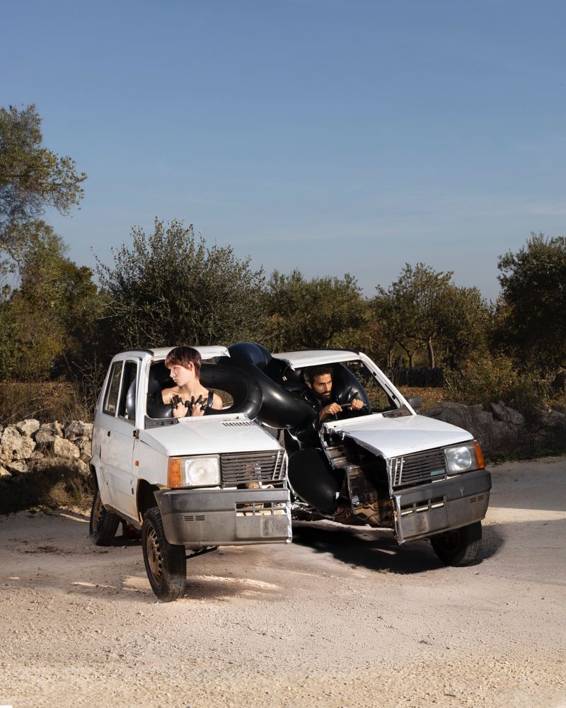 Two vintage white cars merge in a surreal outdoor shot, each with a passenger, connected by a black inflatable tube between them.