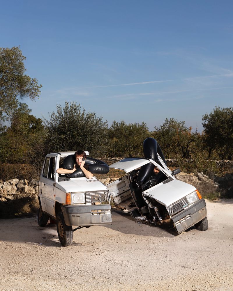 Person in car with phone beside crashed car in an outdoor setting under a blue sky, surrounded by trees, with a dirt road.