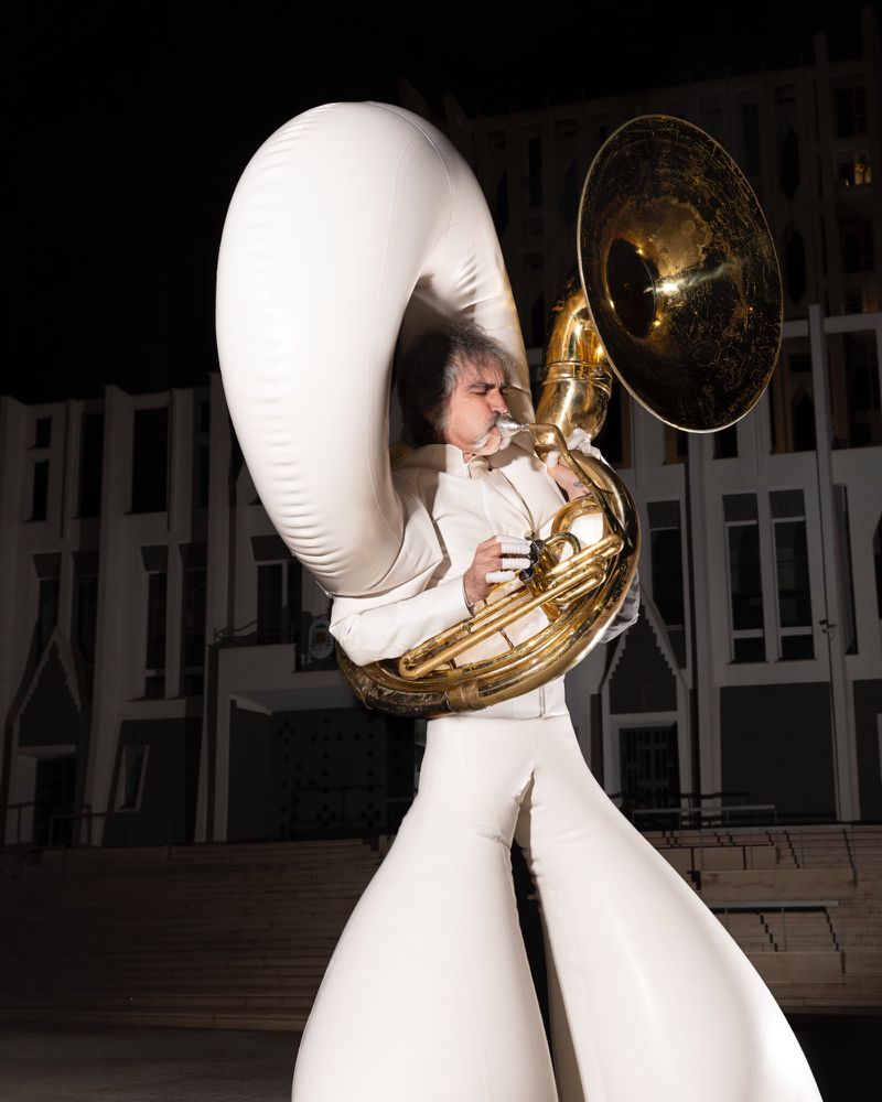 Man in an inflatable white suit plays a sousaphone in front of a modern building at night with dramatic lighting and shadows.