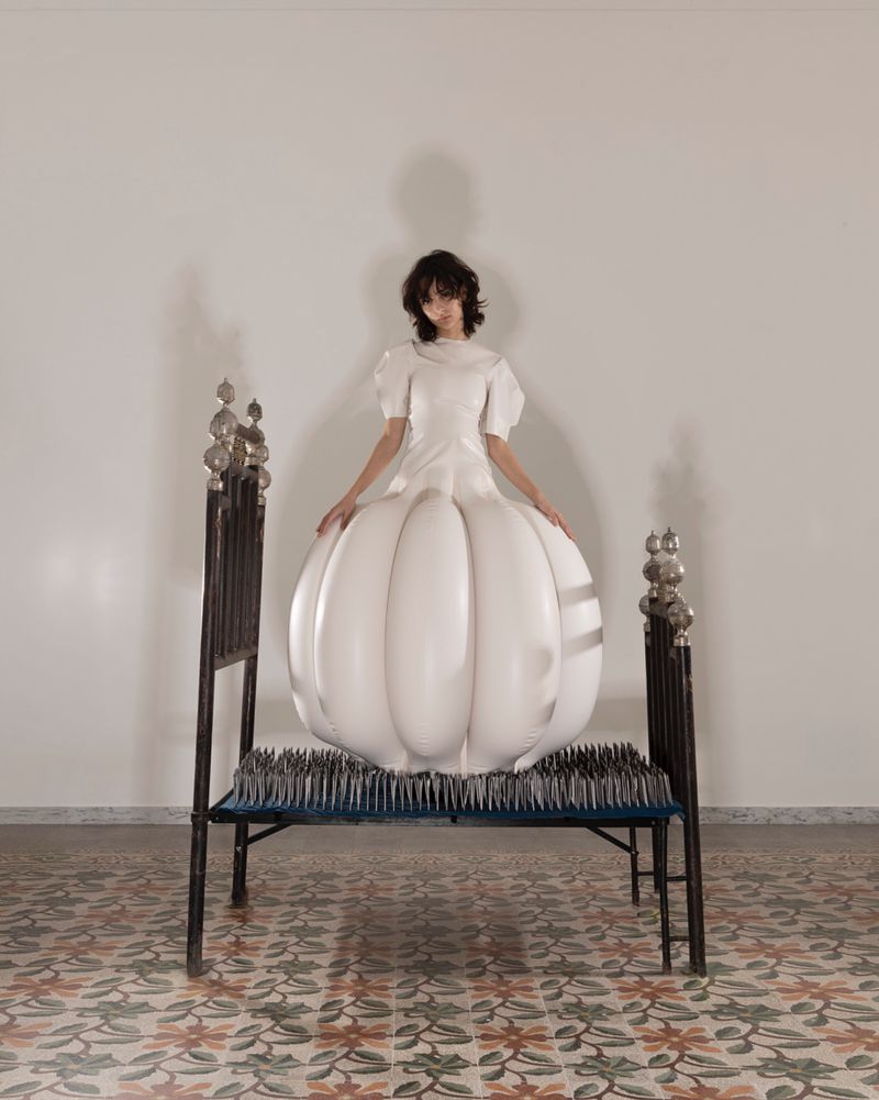 A woman in a white, pumpkin-shaped dress sits on a metal-frame bed covered with spikes in an indoor studio setting with a patterned floor.