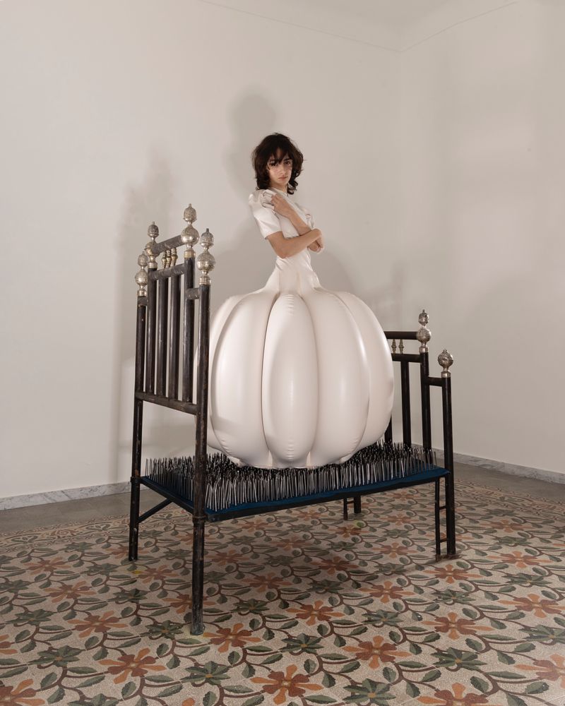 Woman in a white dress with an inflatable skirt on a metal bed frame filled with needles, posing in an indoor setting.