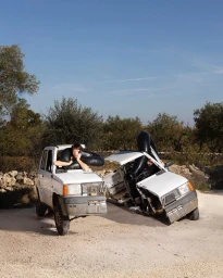 Two people seated inside separate halves of a split white car outdoors on a sandy terrain with trees in the background