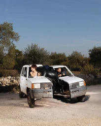 A man and woman sitting inside the halves of a split white car outdoors on a paved road under a clear sky.