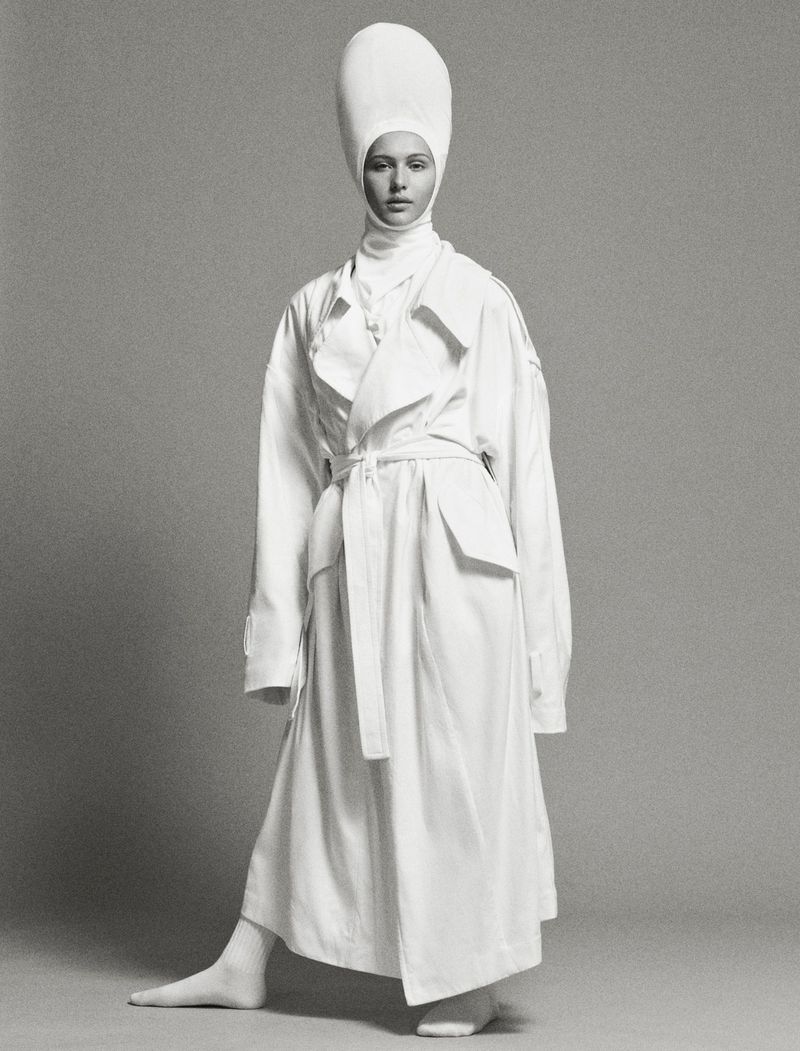 Fashion shot of a woman wearing a white trench coat and a tall white hat against a gray background in a studio setting.