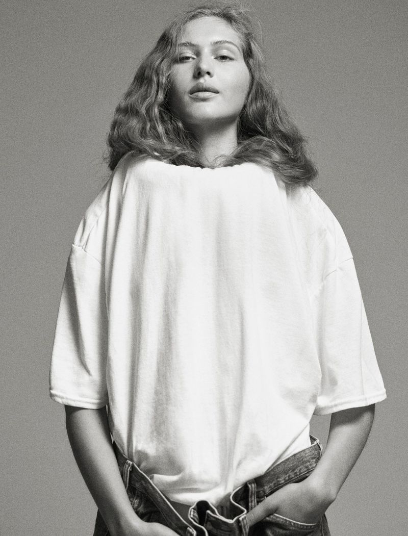 Black-and-white portrait of a young woman with long wavy hair wearing a white T-shirt and denim bottoms against a plain background.