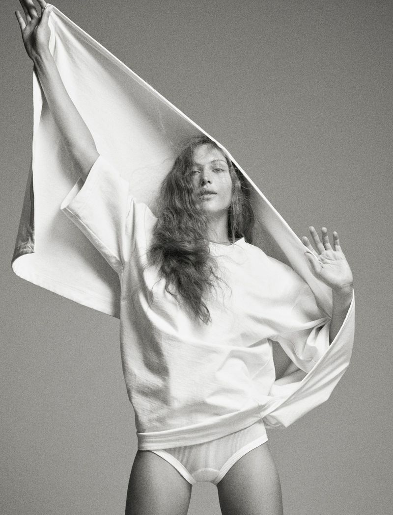 Black and white fashion shot of a young woman in a t-shirt and underwear holding up a white fabric in a studio setting.