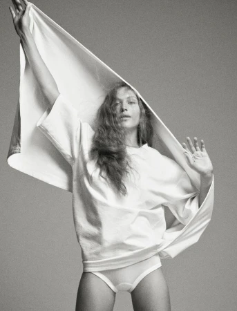 Black and white fashion shot of a young woman in a t-shirt and underwear holding up a white fabric in a studio setting.