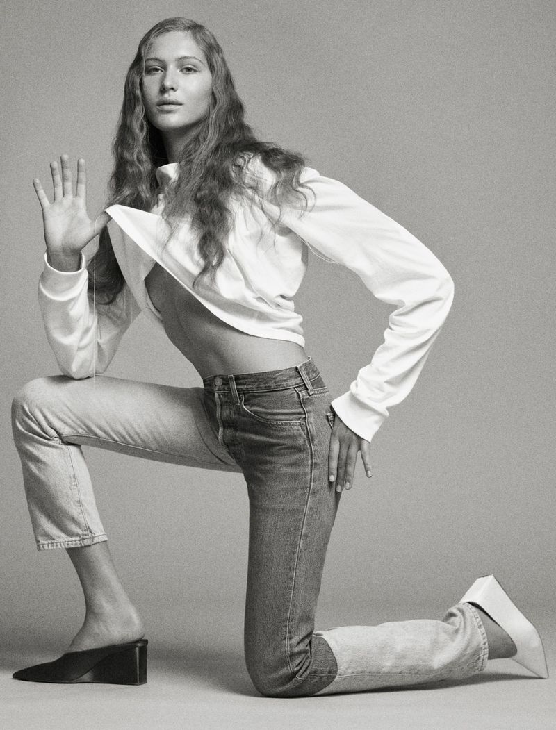 A young woman poses in jeans and a long-sleeved shirt in a studio environment, showcasing fashion and style in a clean, monochrome aesthetic.