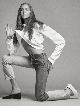 A young woman poses in jeans and a long-sleeved shirt in a studio environment, showcasing fashion and style in a clean, monochrome aesthetic.