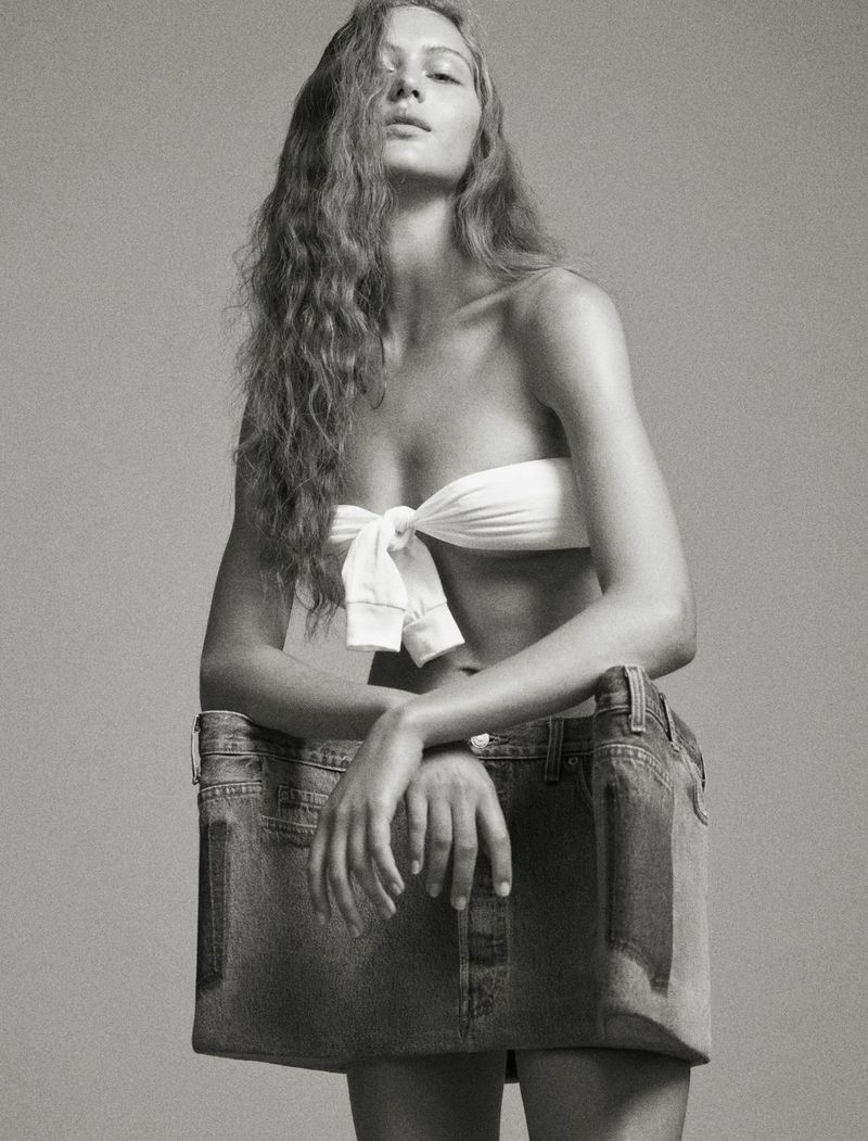 Monochrome studio shot of a woman wearing a white strapless top and a repurposed denim skirt, leaning on the skirt with her arms.