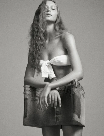 Monochrome studio shot of a woman wearing a white strapless top and a repurposed denim skirt, leaning on the skirt with her arms.