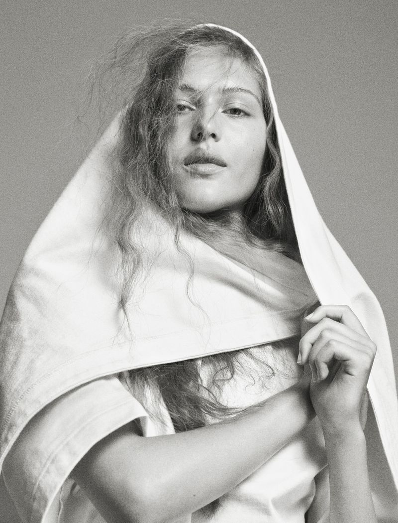 Portrait of a young woman with fair skin and long brown hair, draped in a white cloth, against a solid gray background in a photo studio.