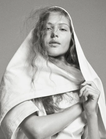 Portrait of a young woman with fair skin and long brown hair, draped in a white cloth, against a solid gray background in a photo studio.